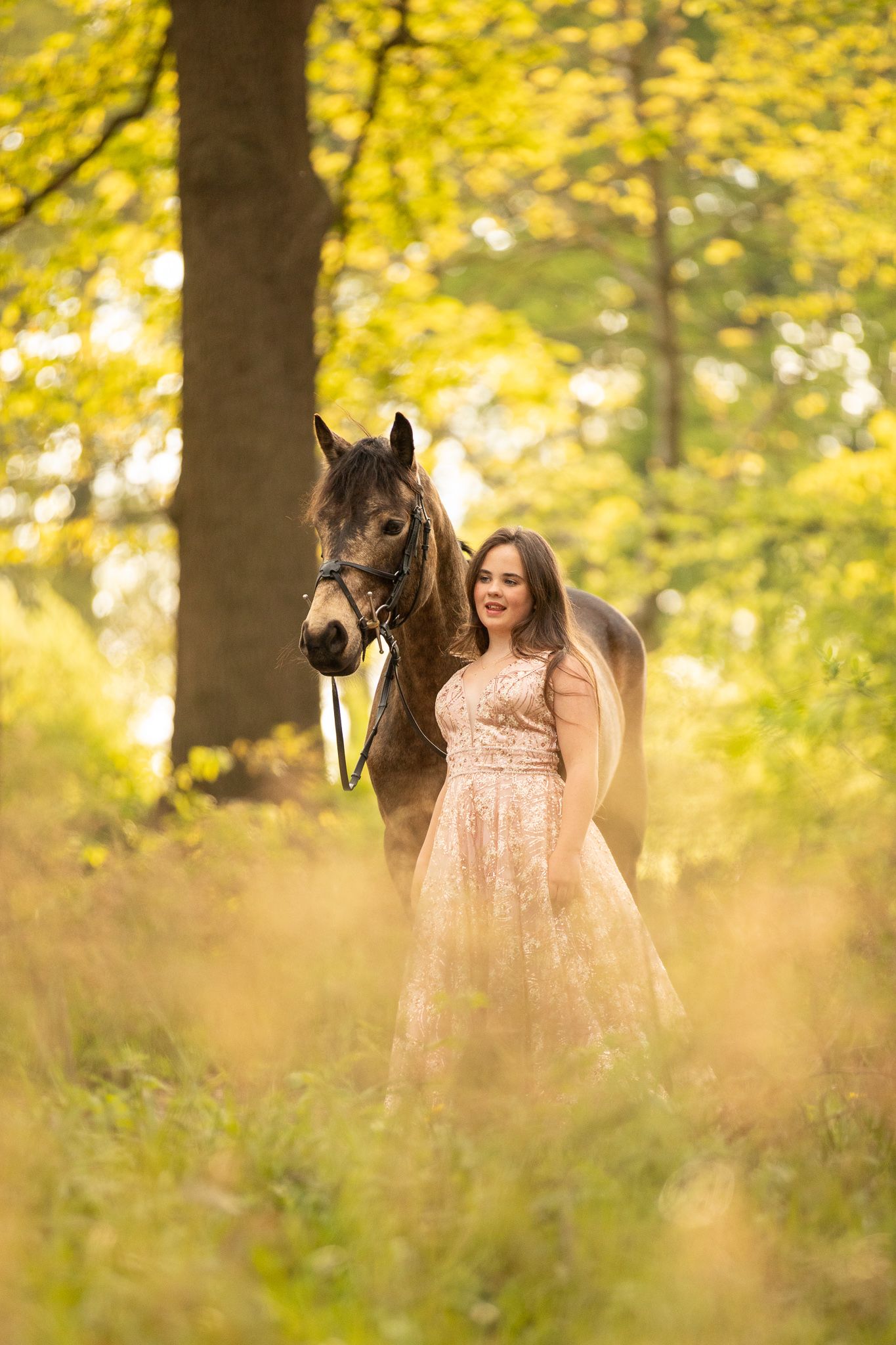 A dreamy autumn scene shows a person in a champagne lace dress standing with a dark brown horse among golden foliage.