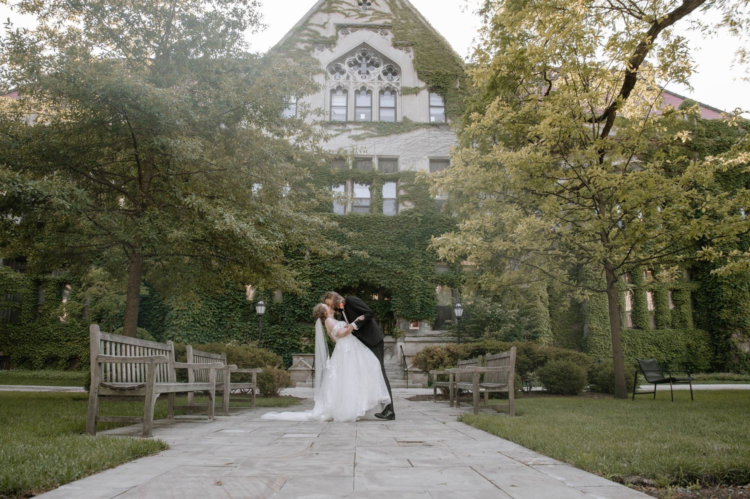 A romantic wedding couple embraces in a serene garden setting with lush greenery and trees.