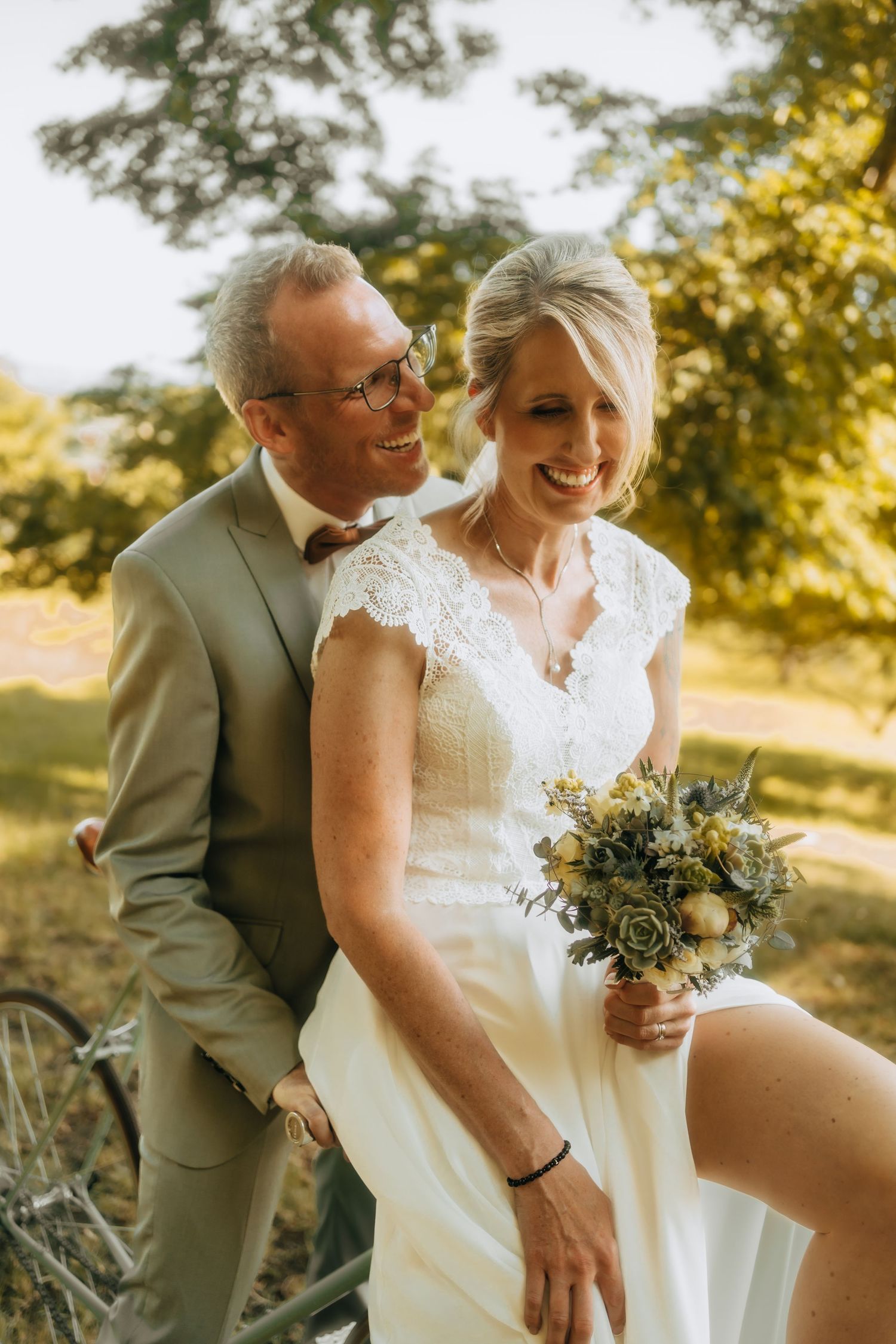 Happy couple laughs together in a sunny outdoor setting wearing wedding attire and holding a lush floral bouquet.