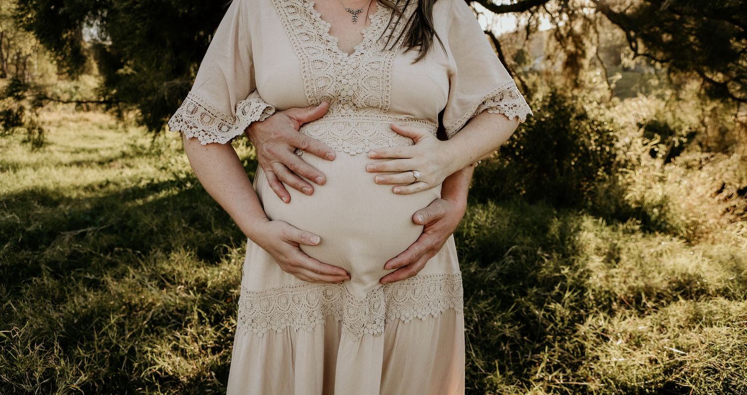 A pregnant person in a flowing cream-colored dress cradles their belly in warm sunlight outdoors.