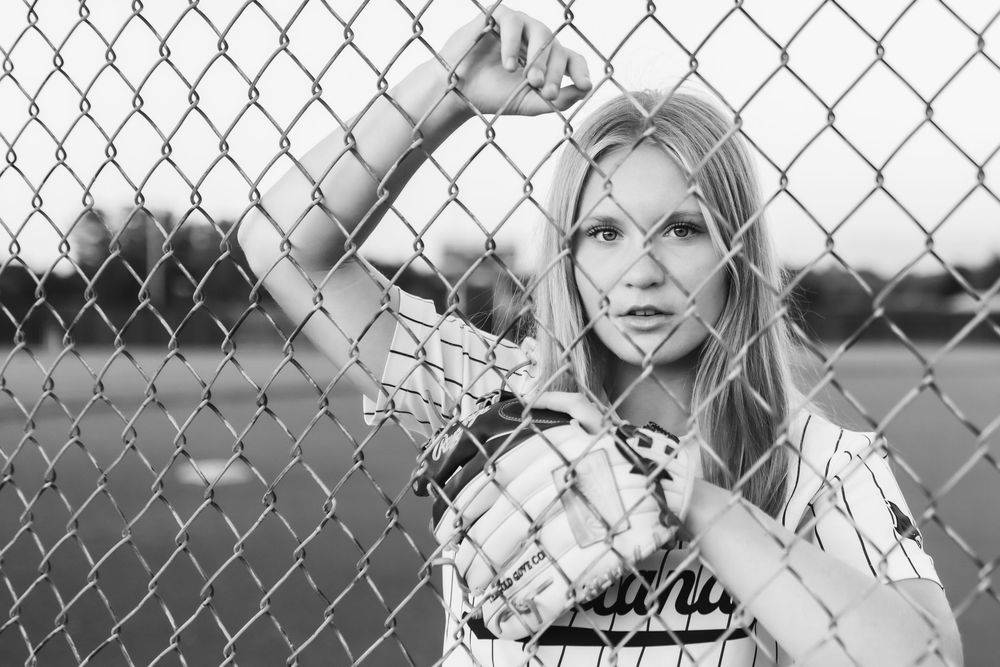 Black and white senior photo of softball player in jersey looking through chain-link fence
