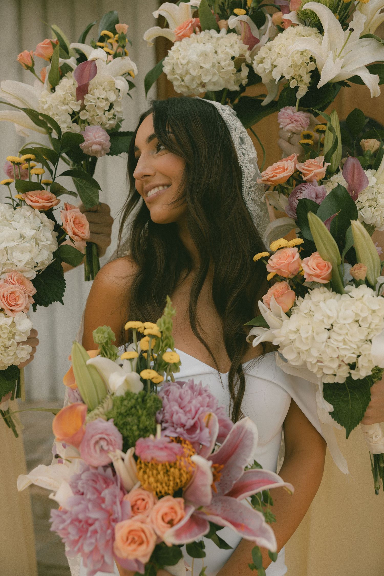 Smiling bride surrounded by colorful floral arch of hydrangeas, roses, and lilies holding pink bouquet.