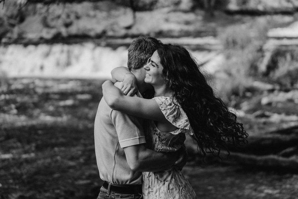Black and white image of engaged couple embracing on a rocky shoreline with roaring falls and forest backdrop