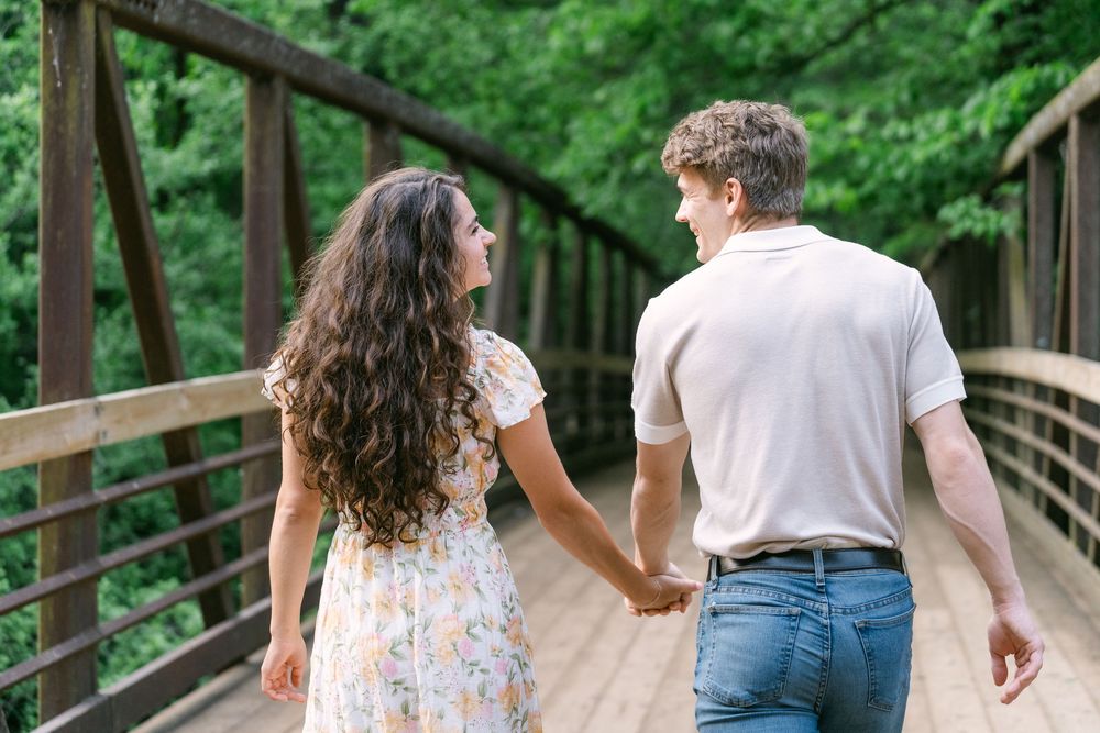 Bride-to-be and fiancé walking away hand-in-hand along a wooded trail during their engagement session