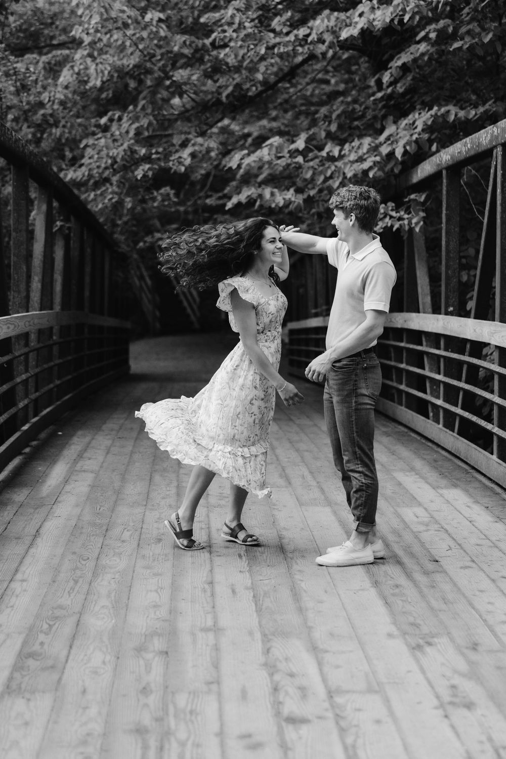 Black and white photo of couple dancing and twirling on a wooden bridge with lush green trees behind them at Willow River State Park