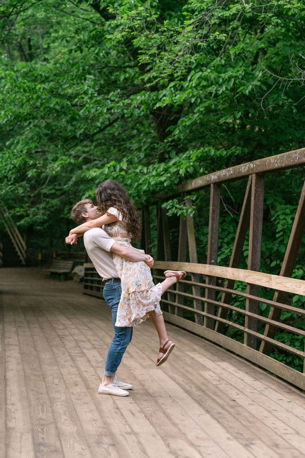Groom-to-be lifting and twirling his fiancée on a wooden bridge surrounded by summer greenery