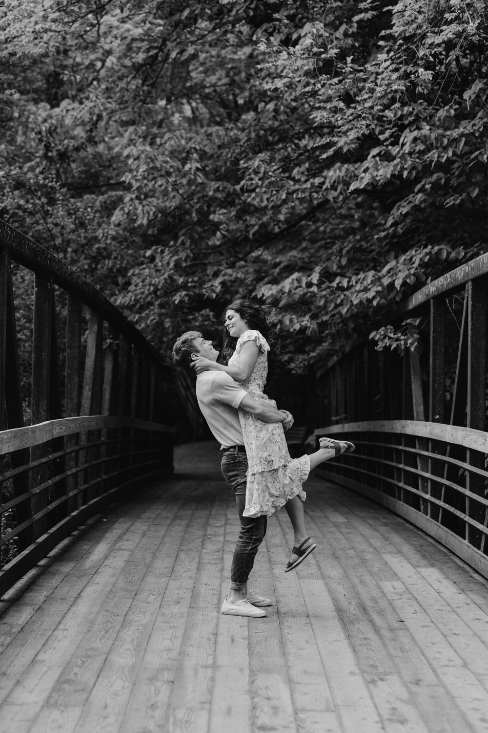 Black and white photo of a lifted kiss on the bridge during a joyful engagement shoot in the woods