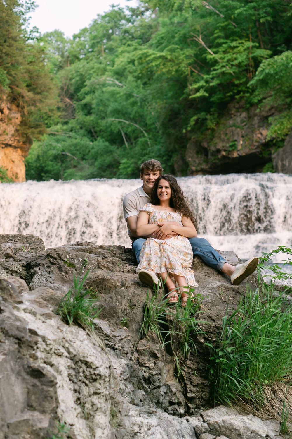 Engaged couple sitting on a rocky ledge with roaring waterfalls and vibrant green trees behind them