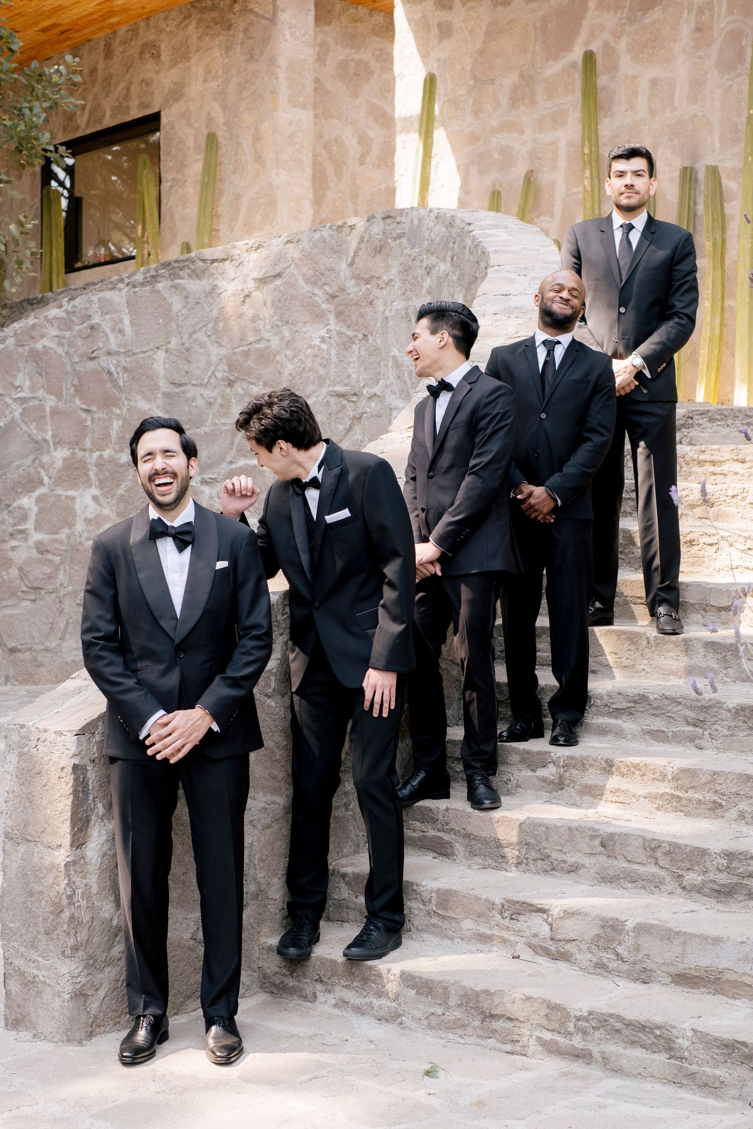 A group of groomsmen in black tuxedos and suits laughing while posing on stone steps at a wedding venue.
