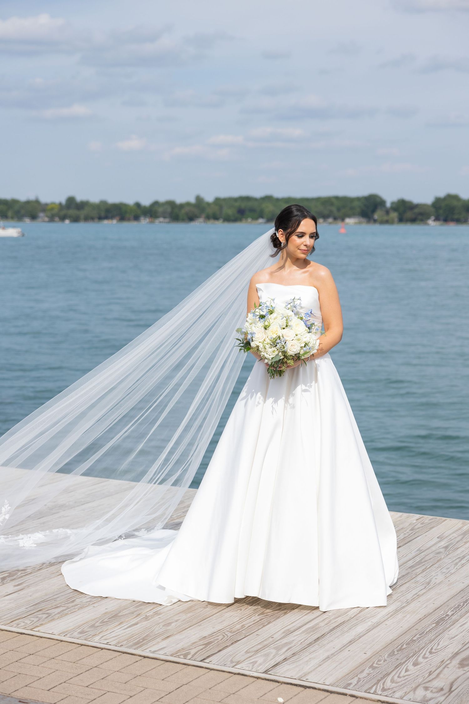 A bride in a white wedding gown poses gracefully on a wooden dock overlooking a serene lake on her special day. Rachel Martinez Photography