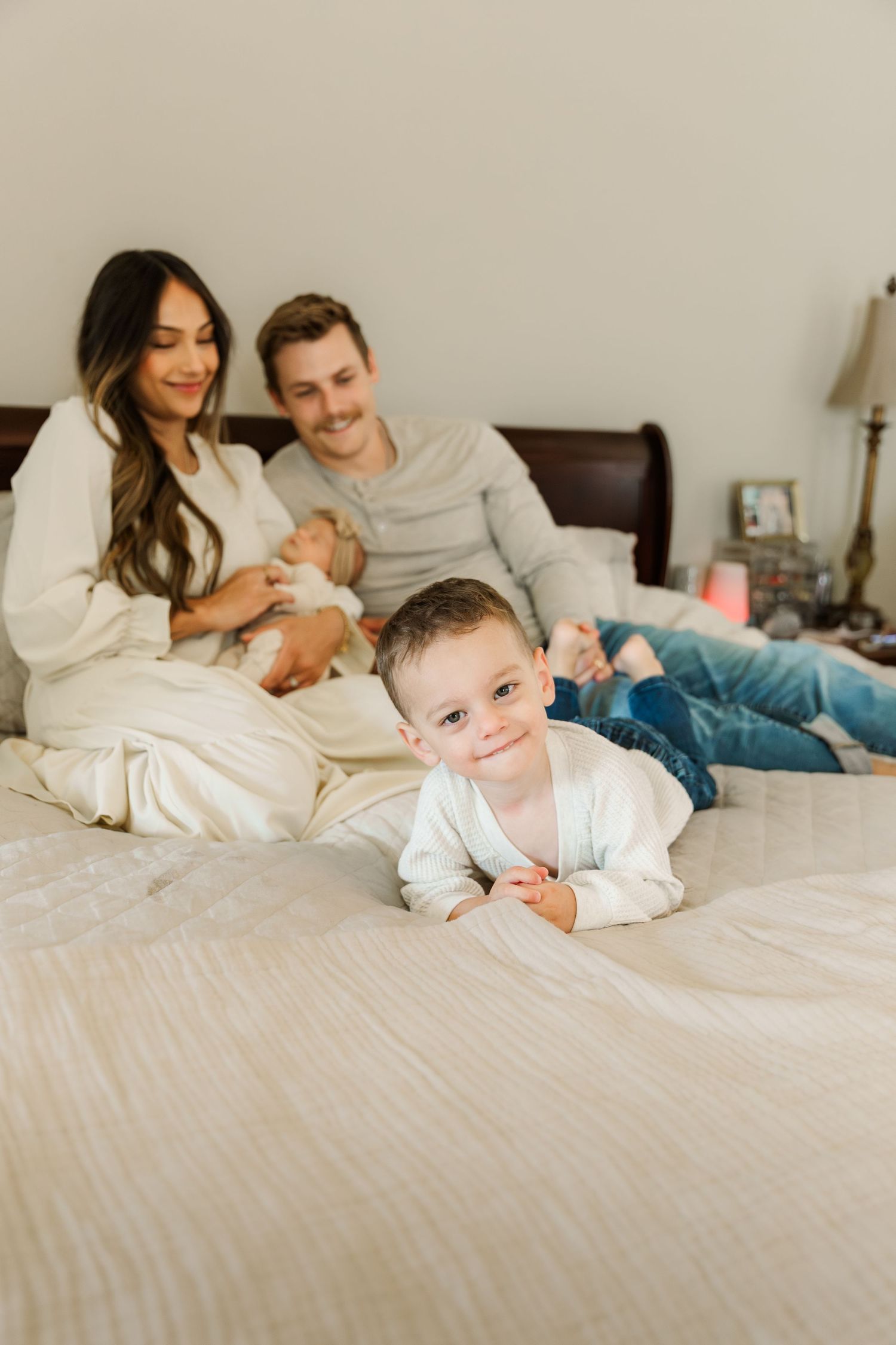 Family lounging together on white bedding while baby plays and crawls in a bright, cozy bedroom setting.