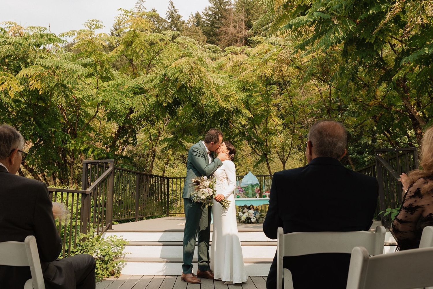 Bride and groom share first kiss on outdoor deck surrounded by lush green trees during ceremony.