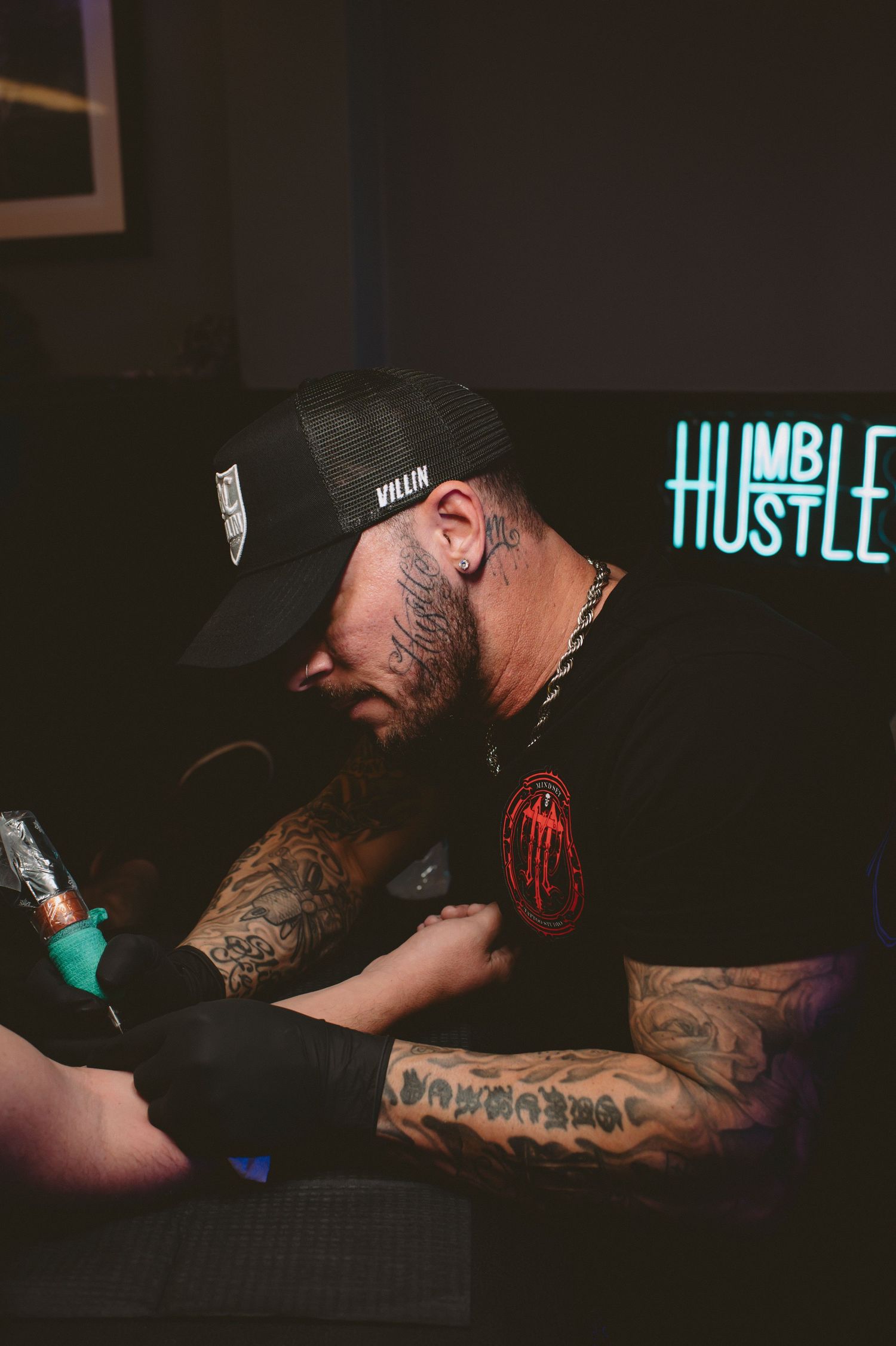 A tattoo artist wearing a black cap works on a client's arm in a dimly lit studio with a neon sign in the background.