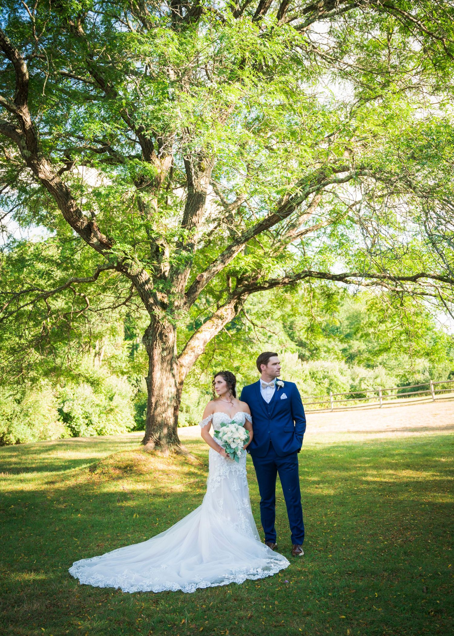 Bride and groom stand beneath a large oak tree on lush green grounds in a summer outdoor wedding portrait.