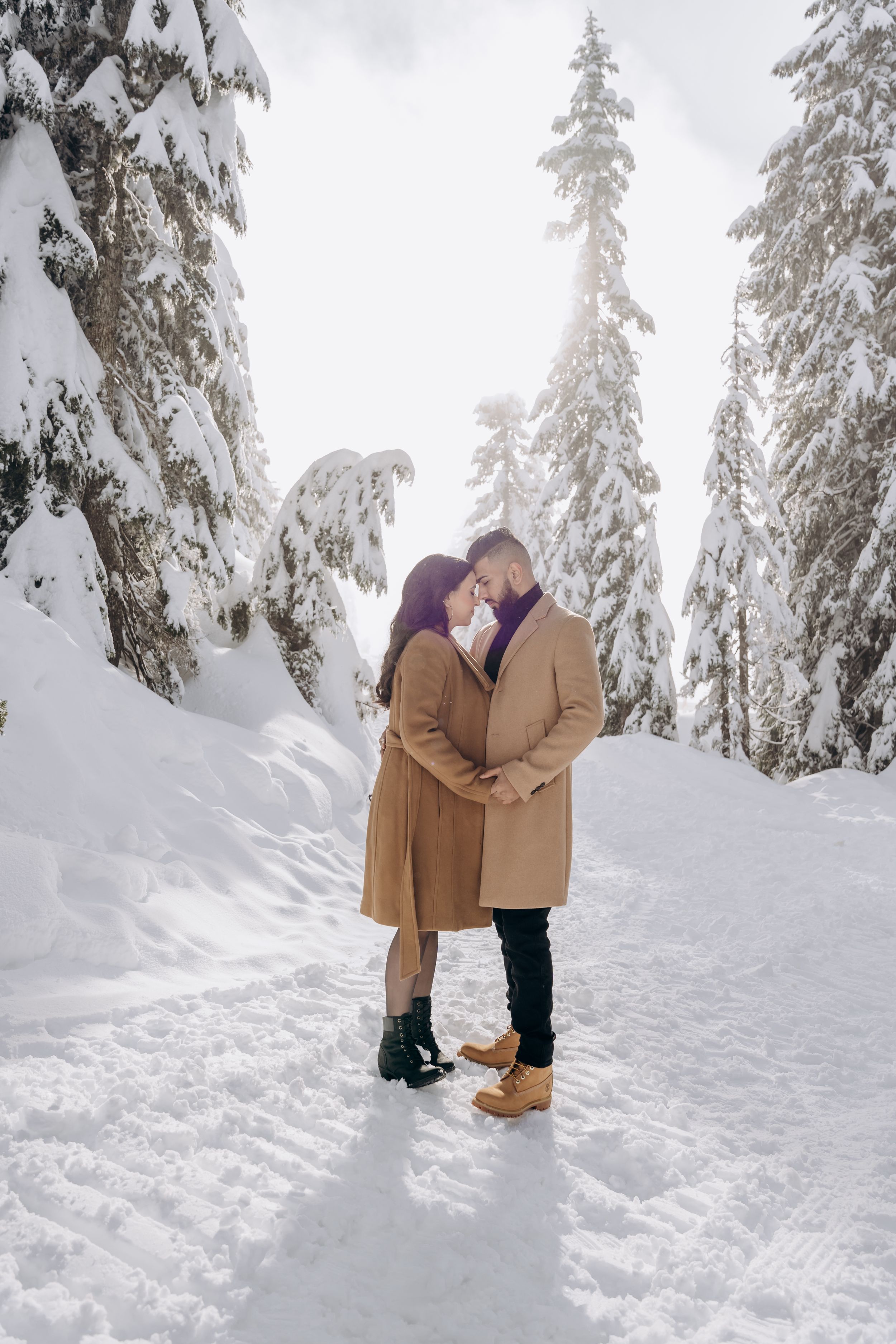 A couple shares intimate winter moments in a snow-covered forest with tall evergreen trees.