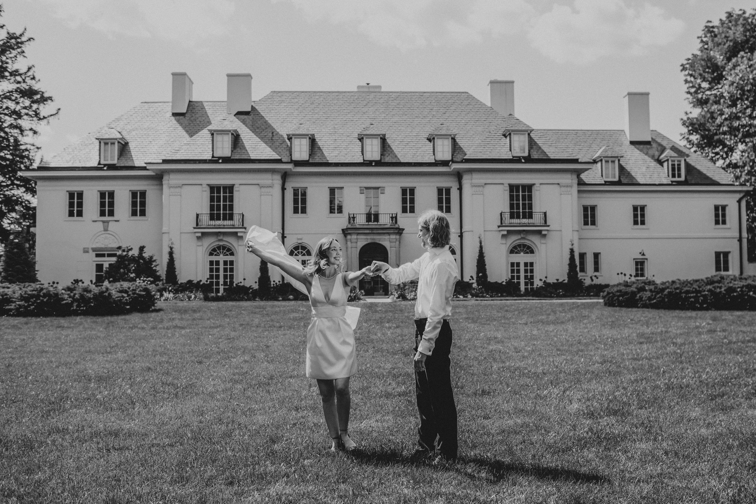 Black and white photo of two people standing on a lawn in front of a grand Georgian-style mansion estate.