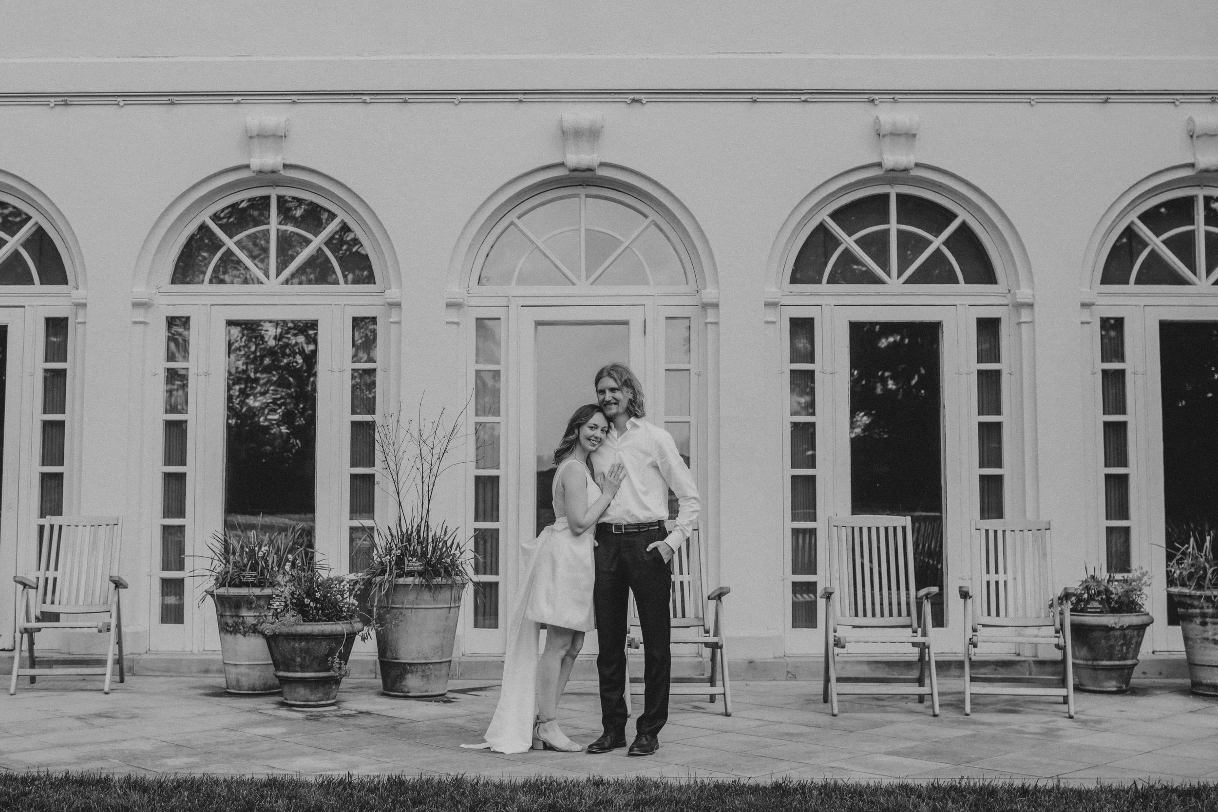 A couple embraces in front of an elegant white building with arched windows and French doors in a black and white photo.