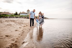 Family of three walking barefoot along sandy beach shoreline near a gazebo on a cloudy day.