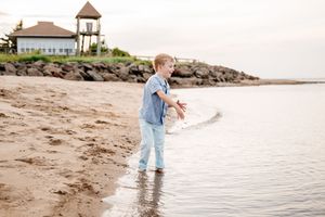 Young boy playing in shallow beach water wearing jeans and a blue shirt near rocky shoreline.