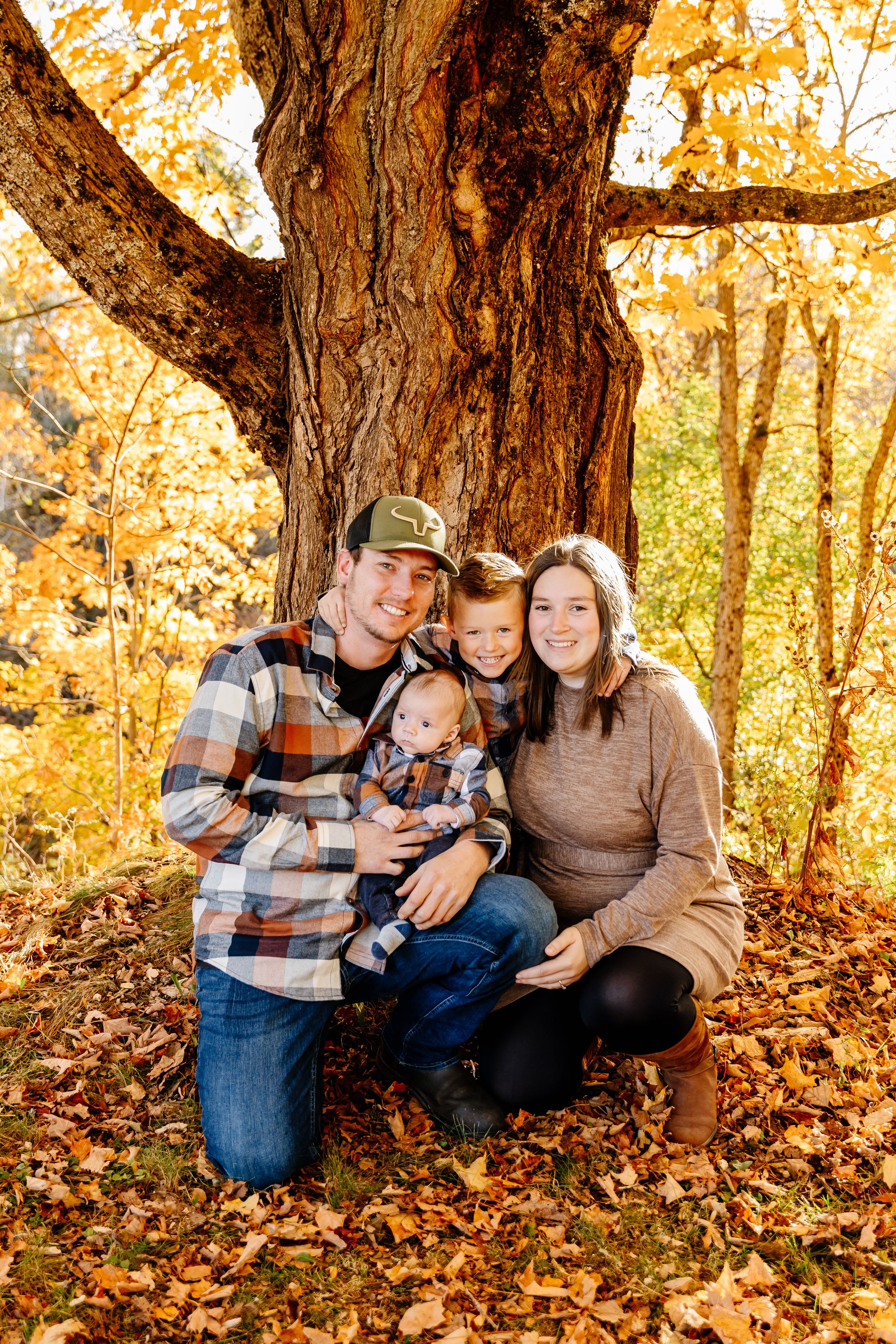 Young family of four posing under a large tree surrounded by vibrant autumn foliage.