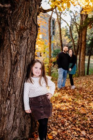 Young girl leaning against a large tree trunk with parents standing behind in fall foliage.