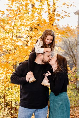 Parents kissing while their young daughter sits on dad's shoulders among golden autumn trees.