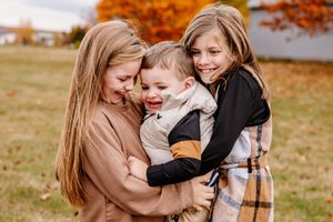 Two sisters hugging their little brother tightly while laughing outdoors in fall foliage.