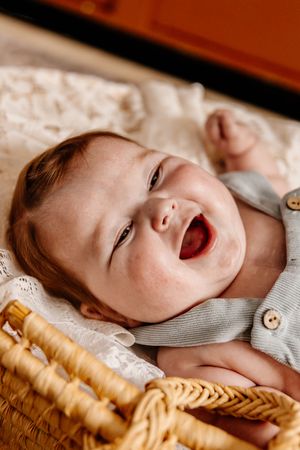Happy laughing baby with freckles lying in a wicker basket wearing a gray button onesie.