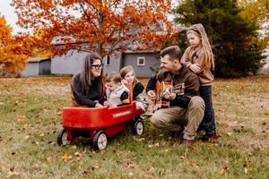 Family of five gathered around a red wagon outdoors among golden and red fall foliage.
