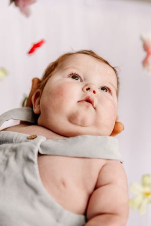 Adorable baby in gray romper gazing upward with chubby cheeks, lying on floral background.