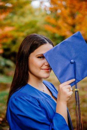 A smiling female graduate in a blue cap and gown playfully covers half her face with her mortarboard. Rogersville
