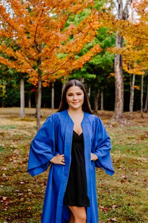 A young woman in a blue graduation gown smiles confidently in front of vibrant autumn-colored trees. Rogersville