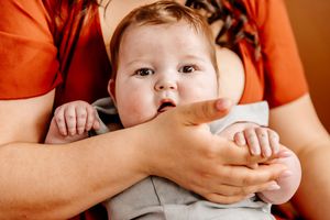 A baby with wide eyes being held securely in an adult's arms, wearing a light gray outfit.