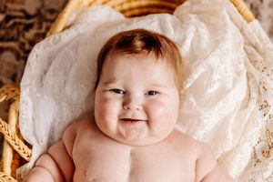 Adorable chubby baby smiling while lying in a wicker basket on a white lace blanket.