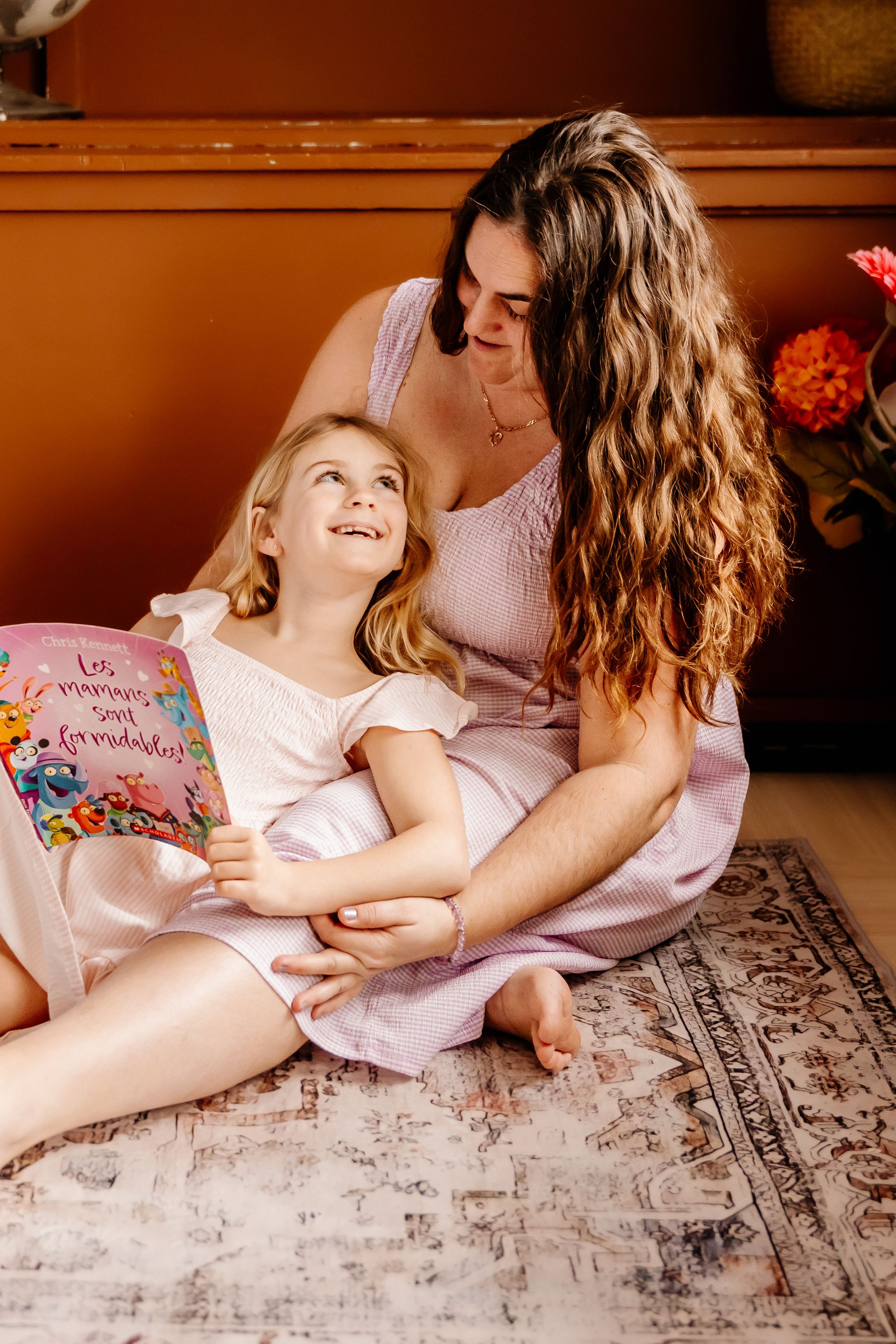A mother and young daughter in pink dresses sit on a rug, sharing a colorful book together.