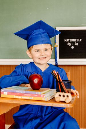 Smiling child in blue graduation cap and gown sits at desk with apple, books, and pencils in front of a chalkboard.