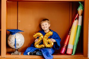 Young boy in blue graduation gown holding gold number 26 balloons, sitting in a shelf with globe and pencils.