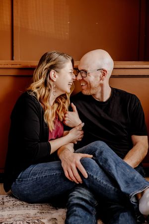 A laughing couple sitting on the floor, embracing and gazing at each other, wearing casual black tops and jeans.