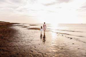 Mother and child walking hand in hand along a serene beach at sunset with reflections on wet sand.