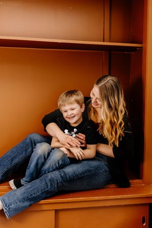 A woman and young boy laugh together while sitting inside an orange wooden cabinet, both wearing black shirts and jeans.