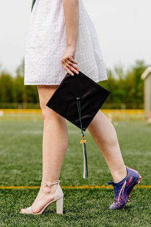 A graduate wearing one heel and one soccer cleat holds a black mortarboard cap on a green field.