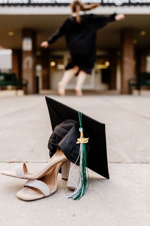A black graduation cap with green tassel and heels on pavement with a jumping graduate in the background.
