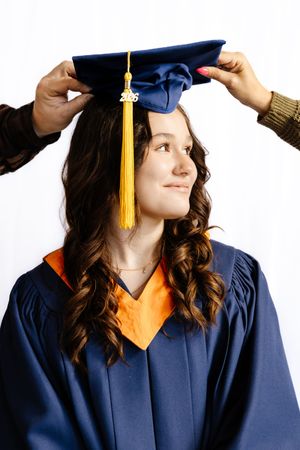 A young woman in a blue graduation gown having her cap placed on her head, with a gold tassel and 2016 charm.