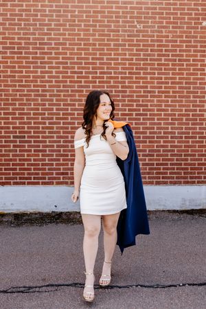 Young woman in white off-shoulder dress holding navy blazer and gold cap, posing in front of red brick wall.