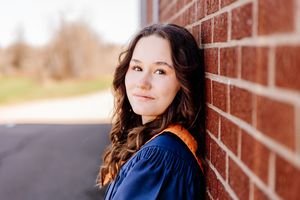 Young woman in blue graduation gown leaning against brick wall, smiling softly outdoors in natural light.