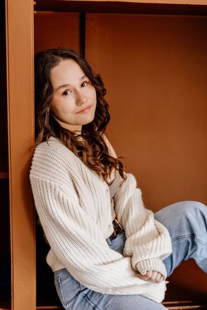 Young woman with curly brown hair in cream knit sweater and jeans sits relaxed against a warm brown wooden shelf.