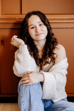 Young woman in white off-shoulder sweater and jeans sitting relaxed against brown cabinet wall.