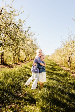 Two young children hugging and smiling in a sunny blossoming apple orchard with green grass and white flowers.