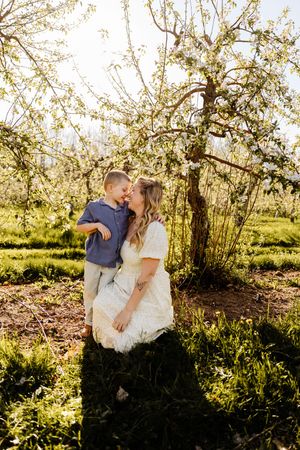 A mother in a white dress holds her young son under a blooming spring tree in a sunlit orchard.