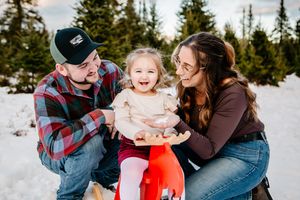 A happy family of three smiles together with their toddler on a red sled in a snowy winter setting.